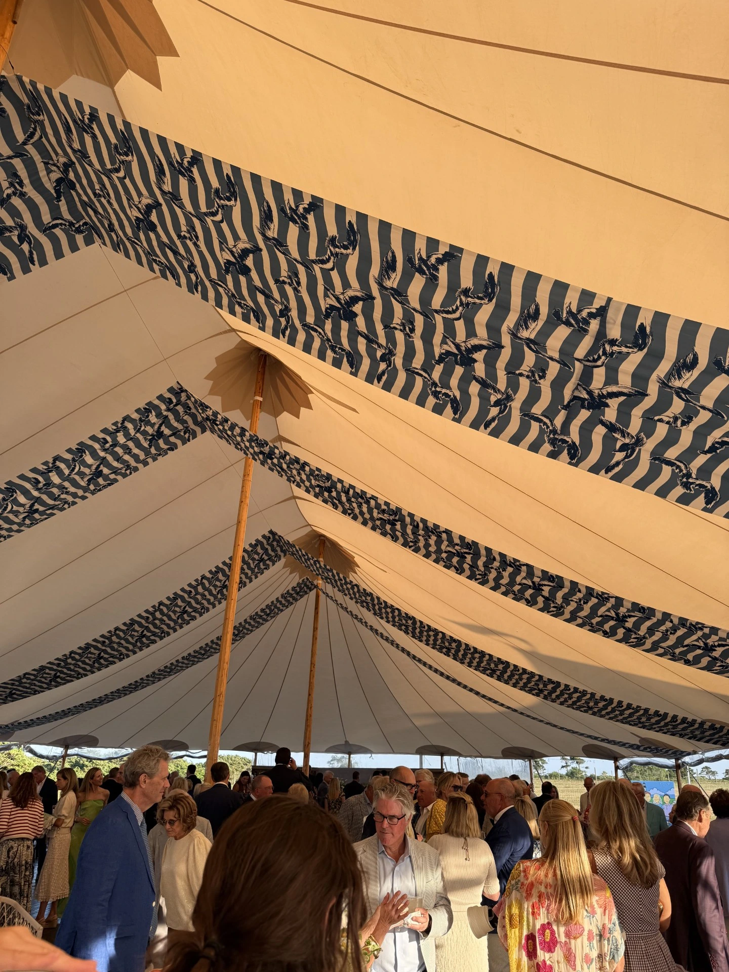 A crowd of people gathered under a large event tent featuring decorative ceiling panels with a blue and white patterned fabric.