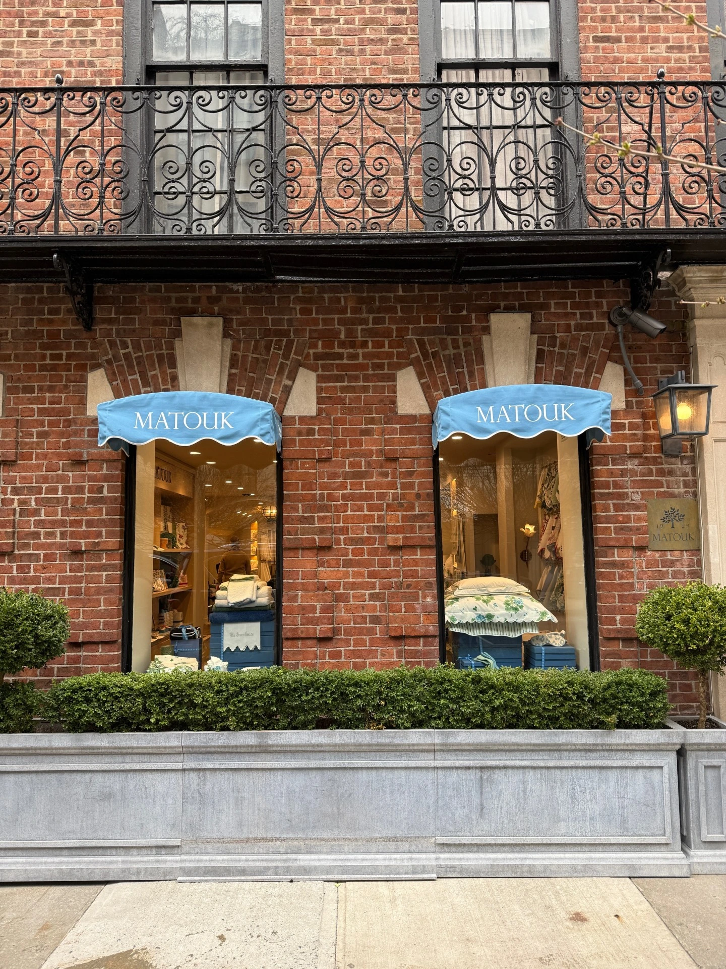 The exterior of a Matouk storefront featuring a classic red brick facade, a wrought-iron balcony, and two bright blue awnings over the display windows.