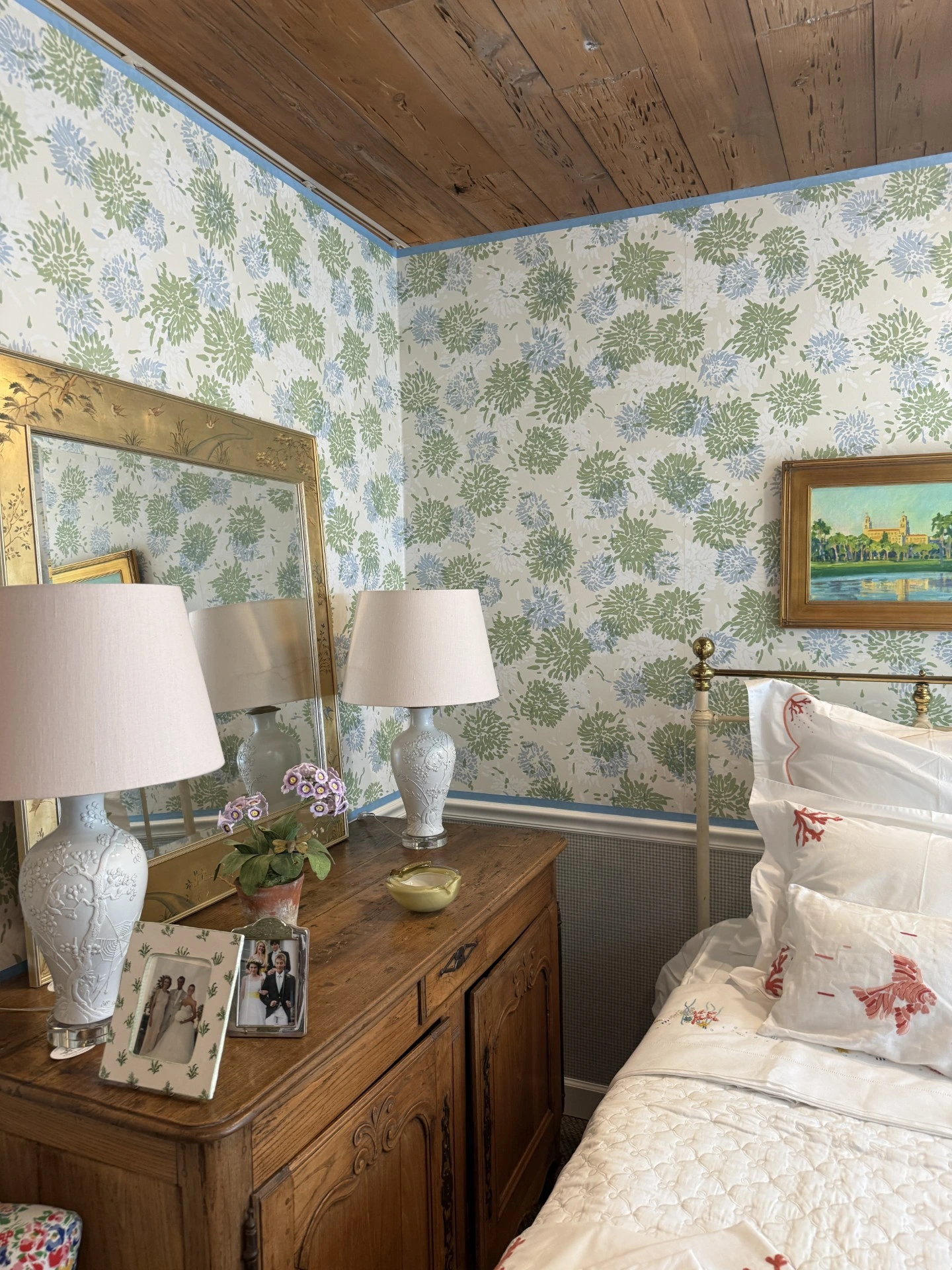 A cozy bedroom interior featuring a wood plank ceiling, green and blue floral wallpaper, a vintage wooden dresser with lamps, and a bed with red-embroidered white linens.