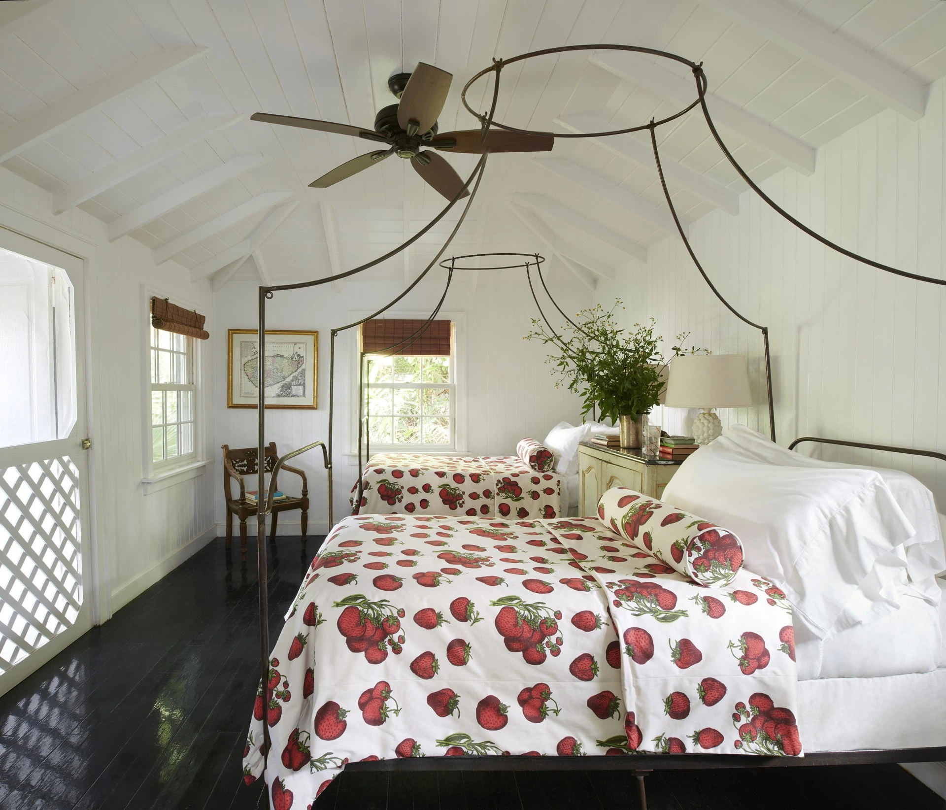 A bright bedroom with a white vaulted ceiling and dark hardwood floors, featuring two metal canopy beds dressed in white bedding with a bold red strawberry print.