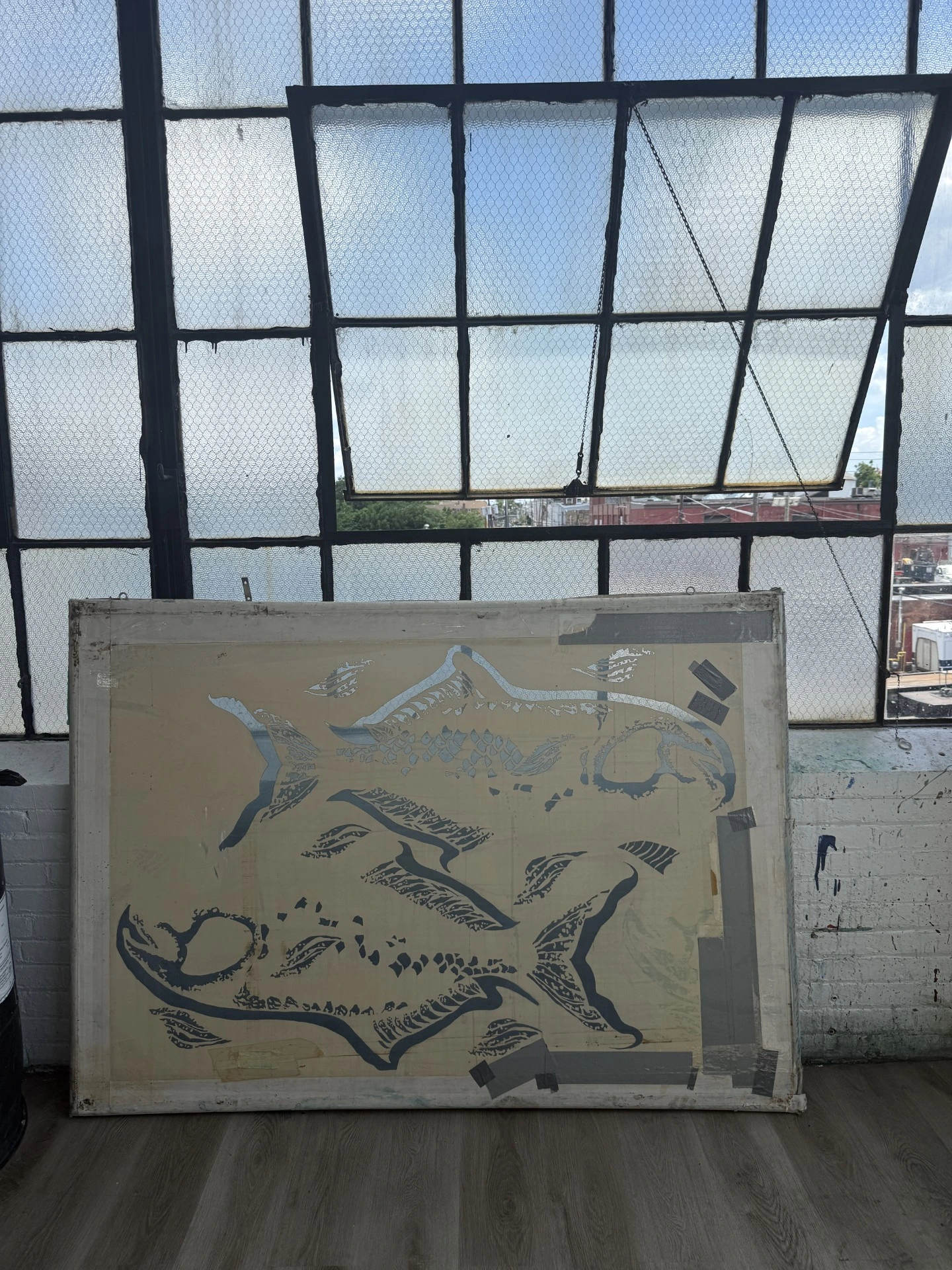 A large screen-printing frame with a stencil of a fish design, leaning against a white brick wall beneath a large industrial window in a studio.