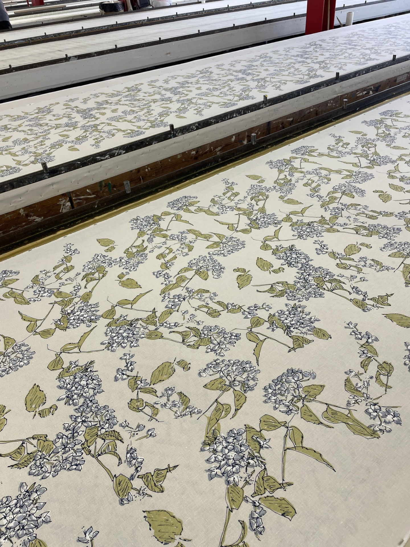 A long expanse of light-colored fabric featuring a green and dark blue floral pattern, laid out on a large production table in a textile facility.