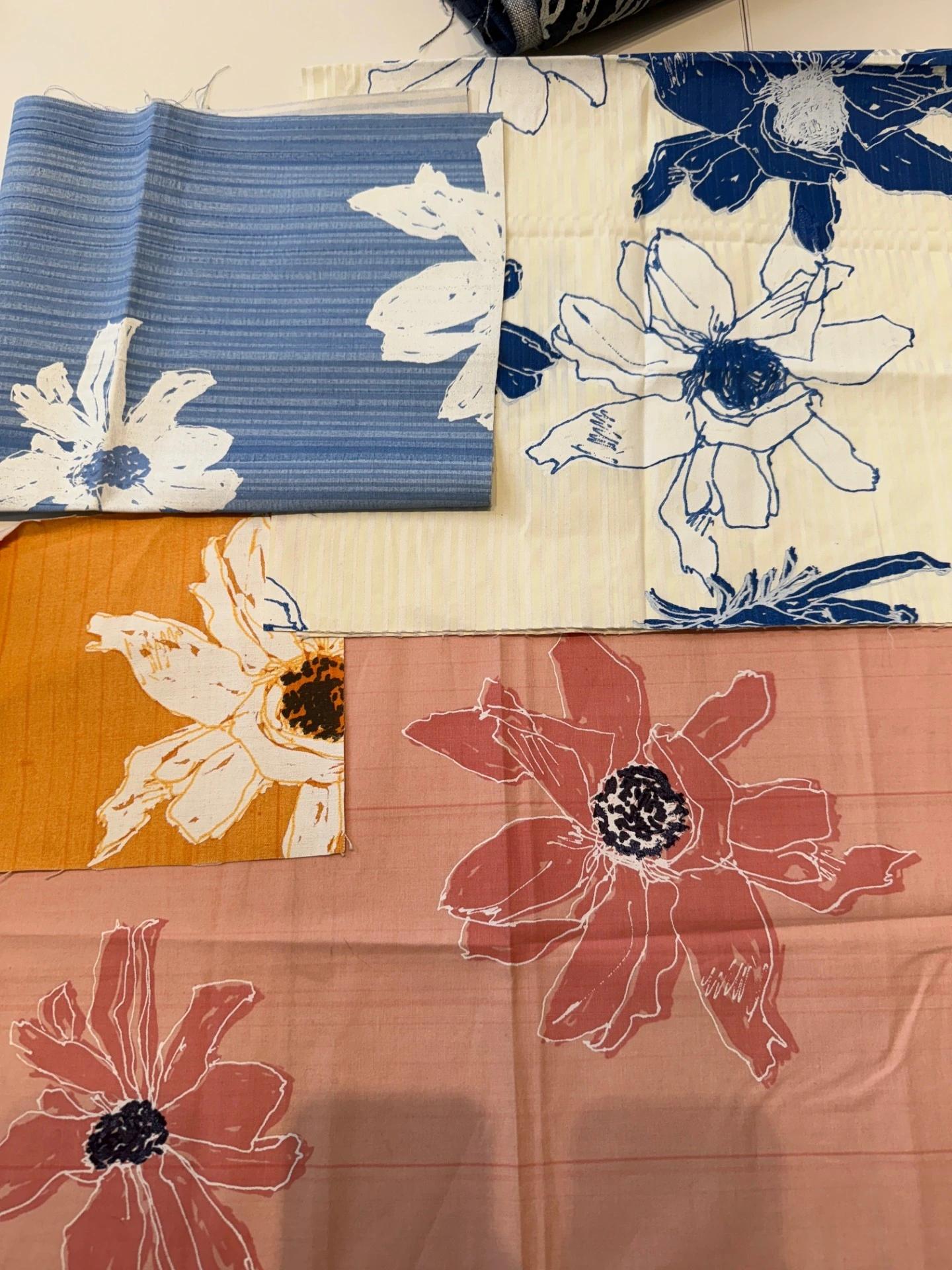 A colorful assortment of textile swatches laid overlapping each other, featuring large, sketchy floral prints in shades of blue, golden yellow, pink, and white.