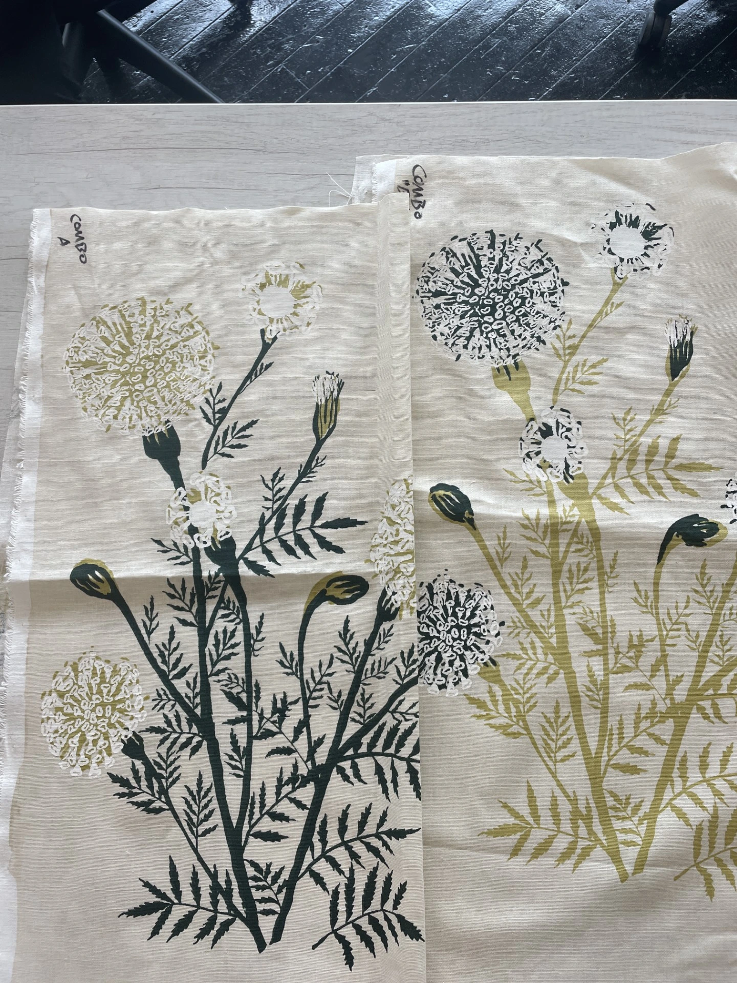 Two fabric swatches laid side-by-side displaying a botanical marigold print in two different colorways: one with dark green stems and light green flowers, and the other entirely in an olive green.